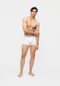 Homme torse nu debout pieds nus en boxer blanc à taille élastiquée "GCDS", posant devant un fond clair uni.