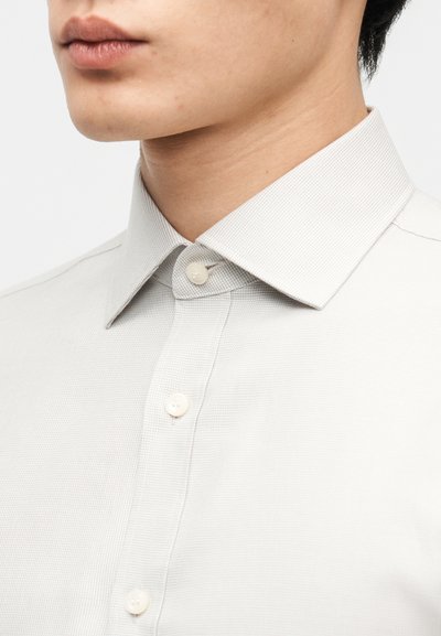 Κοντινή λήψη ενός ατόμου που φορά ένα ανοιχτό γκρι πουκάμισο με υφή, με μυτερό γιακά και τρία ορατά κουμπιά κουμπωμένα.