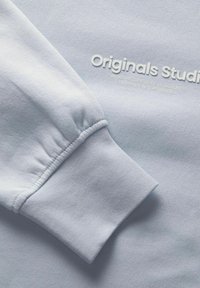 Ljusblå sweatshirt med en åtsittande mudd, prydd med präglad vit text "Originals Studio" och en slät, mjuk textur.