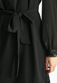 Black dress with a textured fabric, featuring a tied waist and sheer long sleeves. Cuffs adorned with black sequins for added detail.