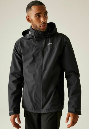 Mężczyzna ubrany w czarną, wodoodporną kurtkę z kapturem i zapinanymi na zamek kieszeniami, patrzący na bok na tle jasnego, jednolitego tła.