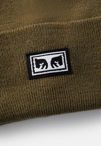 Beanie tricotat de culoare verde olive, cu o textură moale. Dispune de o etichetă rectangulară neagră cu un logo alb ce ilustrează două capete de animale stilizate.