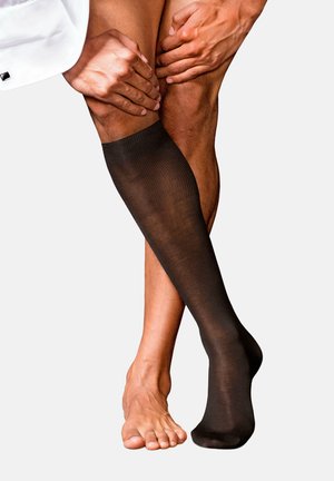 Schwarze Kompressionssocken mit einem gerippten Design, die bis knapp unter das Knie reichen. Die glatte Textur steht im Kontrast zum nackten Fuß und dem Hautton des Beins.