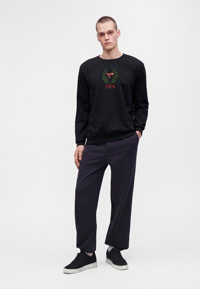 Jeune homme debout, une main dans la poche, portant un sweat-shirt noir avec un logo rouge et vert, un pantalon foncé, des chaussettes blanches et des baskets noires.
