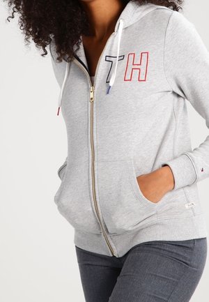 Donna che indossa una felpa con cappuccio grigio chiaro e zip con le iniziali "TH" e le mani nelle tasche anteriori, abbinata a pantaloni grigio scuro.