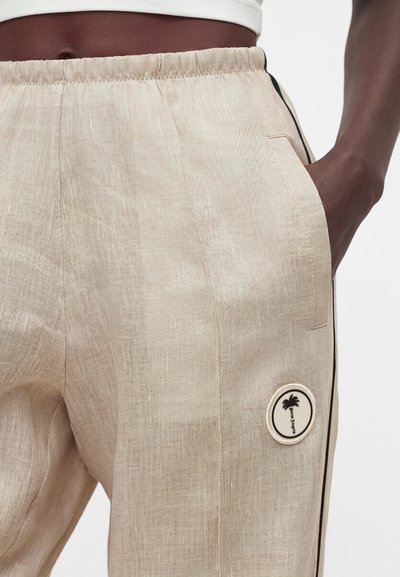 Κοντινό πλάνο ατόμου που φοράει μπεζ λινό παντελόνι με ελαστική μέση, πλαϊνή τσέπη με το χέρι μέσα και στρογγυλό σήμα με φοίνικα.
