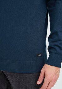 Sötétkék kötött pulóver, bordás mandzsettákkal és aljjal. Az alján egy kis fekete címke található fehér felirattal. Puha tapintású.