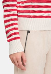 Rot-weiß gestreifter Strickpullover mit geripptem Ärmelbündchen, kombiniert mit weichen, hellbeigen Hosen mit seitlichem Reißverschlussdetail.