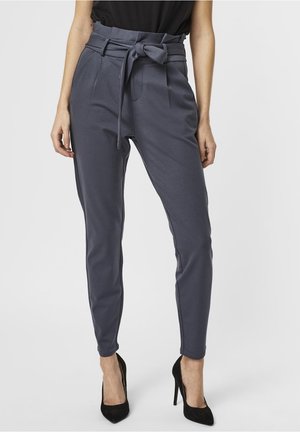 Pantalons gris taille haute avec une ceinture à nœud, design de jambes fuselées et deux poches frontales, associés à des talons hauts noirs.