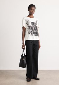 Vrouw in een wit t-shirt met een zwarte panterprint, zwarte wijde broek, met een zwarte handtas, staand voor een effen achtergrond.