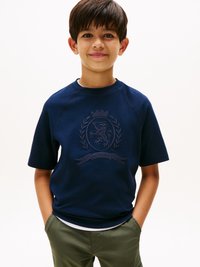 Tricou scurt cu mâneci din material bleumarin, cu un design brodat al unui embleme. Material ușor texturat, croială lejeră, purtat peste o cămașă albă.