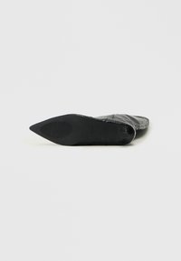 Zapato negro de punta afilada con un upper texturizado, que presenta una suela lisa y un tacón estrecho. El diseño es elegante y moderno.