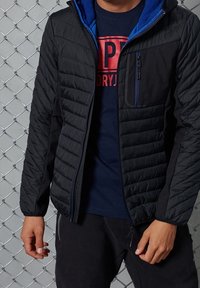 Musta eristetty takki tikatulla kuviolla, sininen vuori, vetoketjullinen rintatasku ja joustavat hihansuut. Päällä tummansininen T-paita.