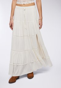 Gonna maxi in morbido tessuto avorio; design a strati con dettagli in pizzo; vita elastica con coulisse; spacco laterale; abbinata a stivaletti marroni.