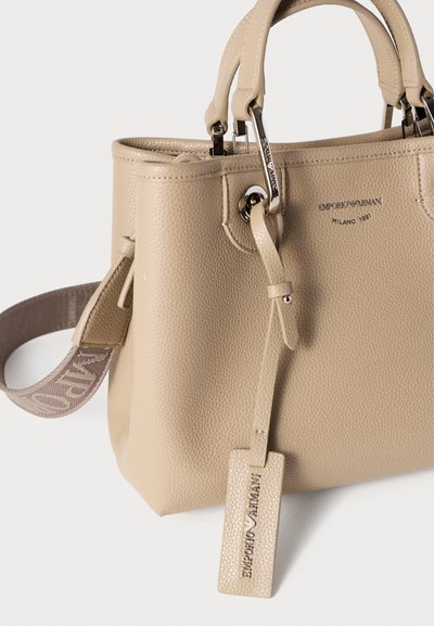 Μπεζ τσάντα από δερματίνη με υφή, με κοντά χερούλια, μεταλλικές λεπτομέρειες και αποσπώμενο λουρί ώμου με μοτίβο λογότυπου. Ετικέτα με το εμπορικό σήμα είναι προσαρτημένη.
