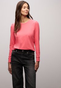 Korallfärgad långärmad tröja med rund halsringning och rynkad detalj vid axeln, tillsammans med svart högmidjade jeans i denim. Mjuk textur.
