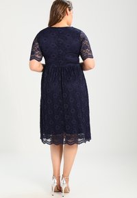 Granatowa sukienka koronkowa z krótkimi rękawami, wzór w kwiaty, dopasowana góra i rozkloszowana spódnica. Posiada długość do kolan i srebrne obcasy.