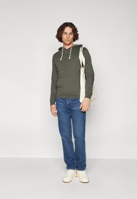 Sudadera con capucha verde oliva y cordones, combinada con jeans de mezclilla azules y zapatillas blancas, junto con un bolso tote de color claro.