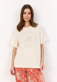 Cream short-sleeve T-shirt with a circular palm tree graphic and text in a tonal print; worn with red patterned pants.