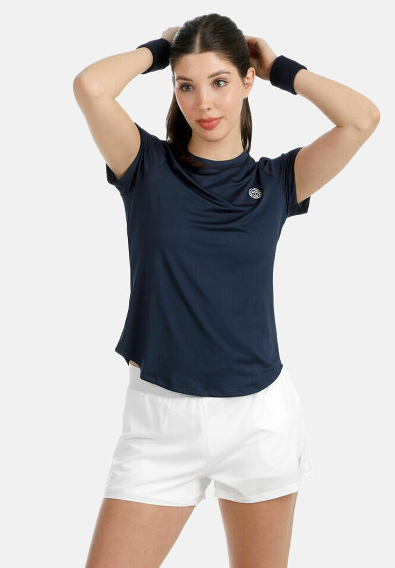 Frau, die ihre Haare richtet, trägt ein marineblaues Sportshirt, weiße Shorts und schwarze Armbänder vor einem schlichten, hellen Hintergrund.