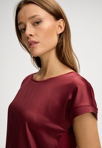 Bordeaux-satin-Oberteil mit Rundhalsausschnitt und kurzen Ärmeln, das eine glatte Textur und einen lockeren Sitz aufweist und sanfte Drapierungen betont.