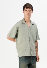Camisa de manga corta de color verde claro con un patrón texturizado, cierre de botones y cuello. Lleva puesta con vaqueros azul oscuro.
