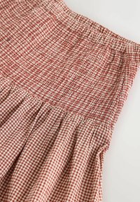 Punane ja valge ruuduline seelik, millel on volditud vöökoht ja volditud detailid. Kerge kangas tekstuuriga viimistlusega.