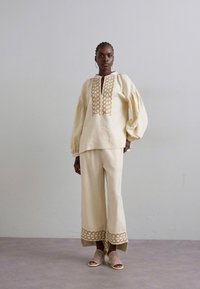 Femme debout dans une tenue couleur crème avec des manches bouffantes, ornementations complexes sur le blouse et le pantalon, portant des talons tressés ouverts.