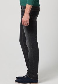 Černé džíny z denimu s přímým střihem a viditelnými švy. Model má na sobě tmavě modré semišové boty. Minimalistický design, bez výrazných vzorů.