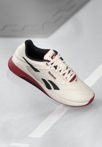 Reebok løbesko med en cremefarvet overdel, sorte accenter og rød sål; tekstureret design med snørebånd og logo på tungen.