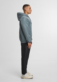 Kapucňová bunda v tlumené modro-zelené barvě s hladkým povrchem, s přední kapsou a elastickými manžetami, doplněná o černé kalhoty a světlé tenisky.