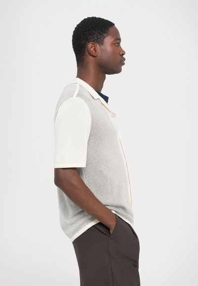 Short-sleeve knit shirt in light beige and cream, featuring a buttoned front and breathable texture. Styled with dark trousers.