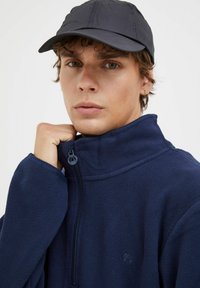 Giacca in pile blu navy con colletto alto e zip; texture morbida; indossata con un berretto scuro; presenta un logo vicino al petto.