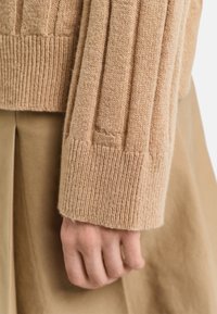 Primo piano di una mano appoggiata di lato, che indossa un maglione a coste beige e pantaloni a pieghe color beige.