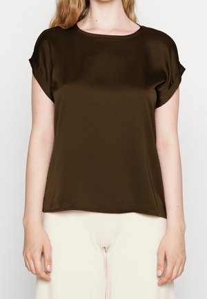 Vrouw met een bruin kortärmelig topje en lichtbeige broek, getoond van schouders tot heupen tegen een effen achtergrond.