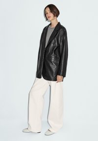 Zwarte leren blazer met een ontspannen pasvorm, twee voorkoeken en knoopsluiting, gecombineerd met losse witte broeken en lichte schoenen.