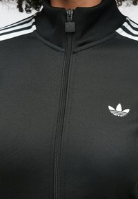 Fekete, texturált anyagból készült cipzáras dzseki, három fehér csíkkal a vállakon és egy kis fehér logóval a bal mellkason.