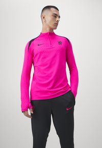 Rosa Sportshirt mit langen Ärmeln und Reißverschlusskragen, strukturiertem Stoff, schwarzen Akzenten und Logo. Kombiniert mit schwarzen Hosen, die ein kleines Logo aufweisen.