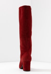 Czerwone zamszowe buty za kolano z grubym obcasem i bocznym zamkiem. Charakteryzują się gładką teksturą i wyraźnym kształtem, zaprojektowane dla dopasowanego wyglądu.