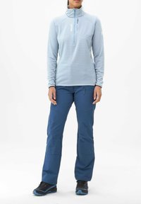 Pull bleu clair avec une fermeture éclair quart, en tissu texturé, et un logo bleu, associé à un pantalon bleu avec des poches et une coupe ajustée. Chaussures noires.
