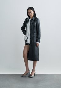 Schwarzer Ledermantel mit Knöpfen und Taschen, über einem weißen Hemd mit Knopfleiste getragen, kombiniert mit glänzenden silbernen Sandalen mit Blockabsatz.