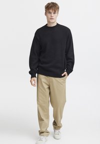 Suéter negro de punto con escote redondo, combinado con pantalones beige de corte holgado y zapatillas blancas, sobre un fondo blanco liso.