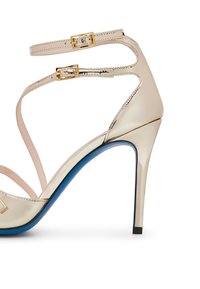 Sandal con tacco alto beige con finitura lucida in vernice, doppi cinturini alla caviglia, fibbie dorate e dettaglio sulla suola blu; punta affusolata e tacco stretto.