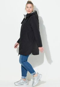 Schwarze Kapuzenjacke mit Reißverschluss und seitlichen Taschen. Graue Innenauskleidung, abgerundeter Saum, kombiniert mit blauen Jeans und silbernen Sportschuhen.