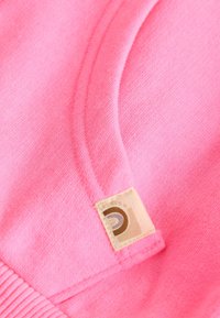 Rožinės audinio detalės su išsamiu siuvimu ir mažu smėlio spalvos ženkliuku, kuriame yra rusvos spalvos kreivė, šalia siūlės.