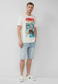 T-shirt blanche avec imprimé graphique représentant un requin et des bateaux. Short en denim bleu clair. Baskets blanches décontractées avec des accents gris.