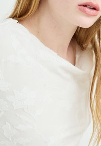 Hvit topp med et teksturert blomsterdesign, med rund hals og mykt stoff. Bakgrunnen er nøytral, som fremhever detaljene i plagget.