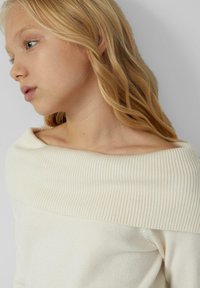 Jonge blonde vrouw met golvend haar, gekleed in een crème ribbeltricot trui met off-shoulder, kijkt naar links tegen een effen achtergrond.