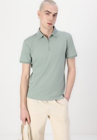 Polo majice mint zeleno zadrženim ovratnikom na zips, kratkim rukavima in mehko teksturo. Obute z svetlo bež hlačami in drži torbo. 