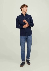 Jovem a ajustar a manga, vestindo uma camisa azul marinho de botões, jeans azuis com bainhas e ténis pretos contra um fundo claro liso.